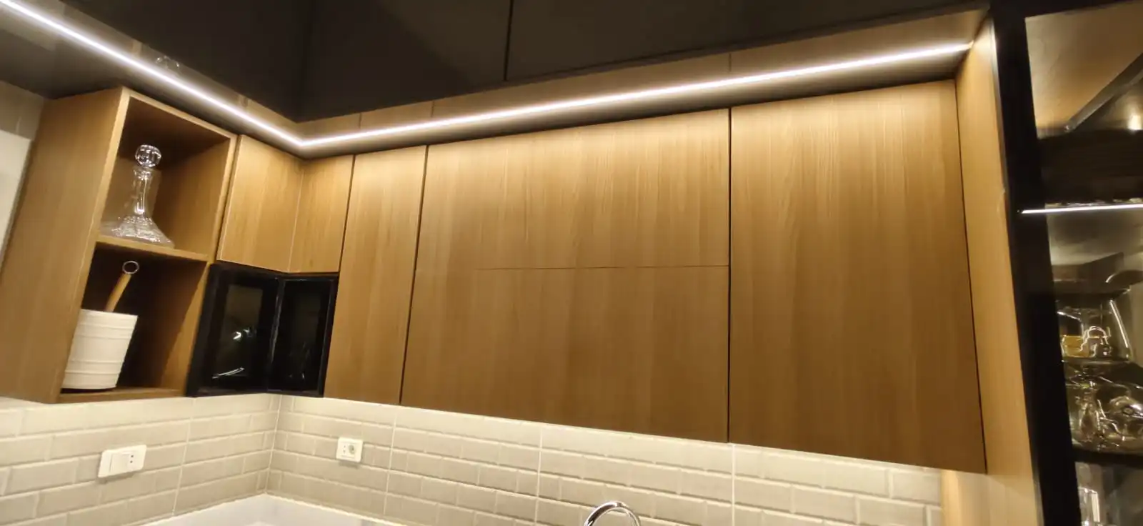 دواليب مطبخ علوية بلون البلوط مع شريط إضاءة LED في أعلاها فوق حائط من البلاط الأبيض